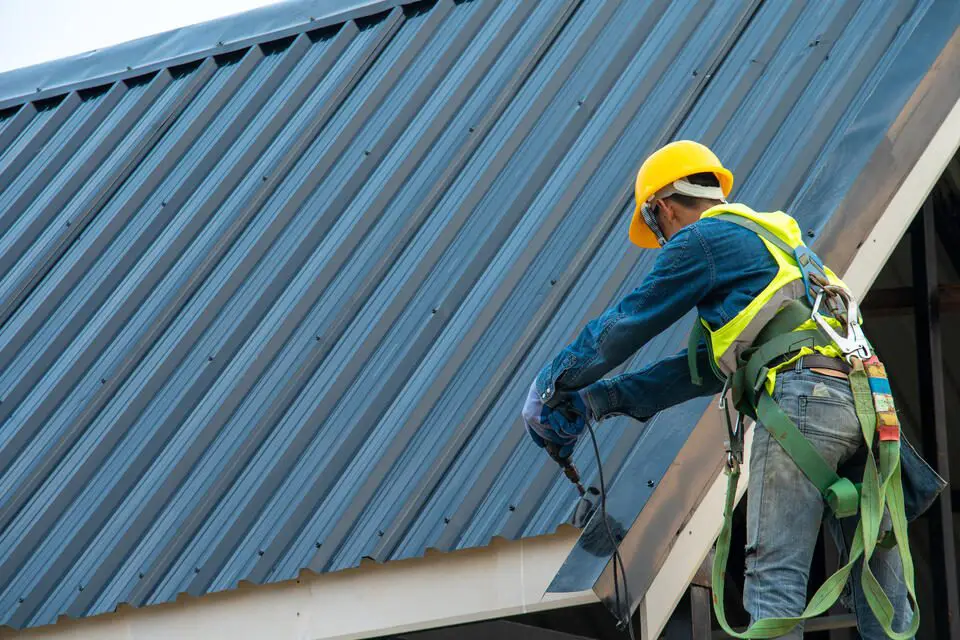 metal roof cost-roofer wearing safety gear installing metal roofing panels on a sloped roof