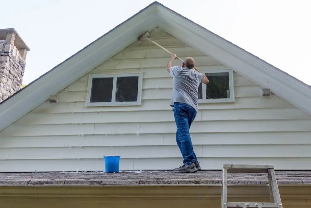 Man stands on porch roof to wash house with brush on extension