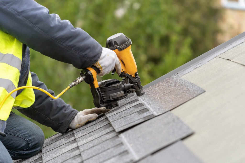 Unrecognizable roofer worker in uniform work wear using air or pneumatic nail gun and installing asphalt or bitumen tile on top of the roof under construction house