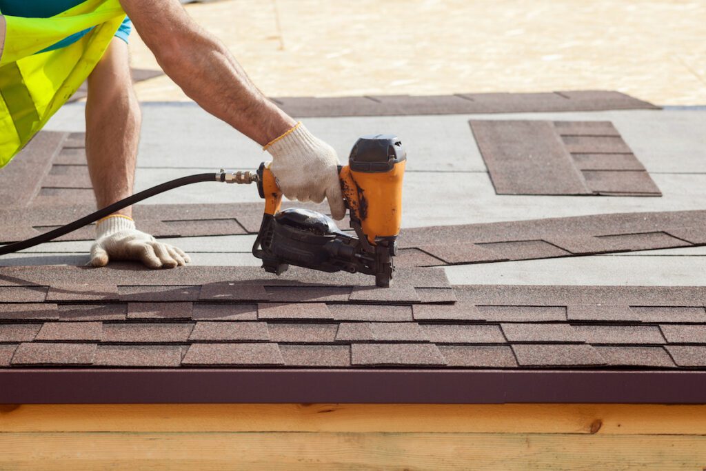 Construction worker putting the asphalt roofing (shingles) with nail gun on a new frame house