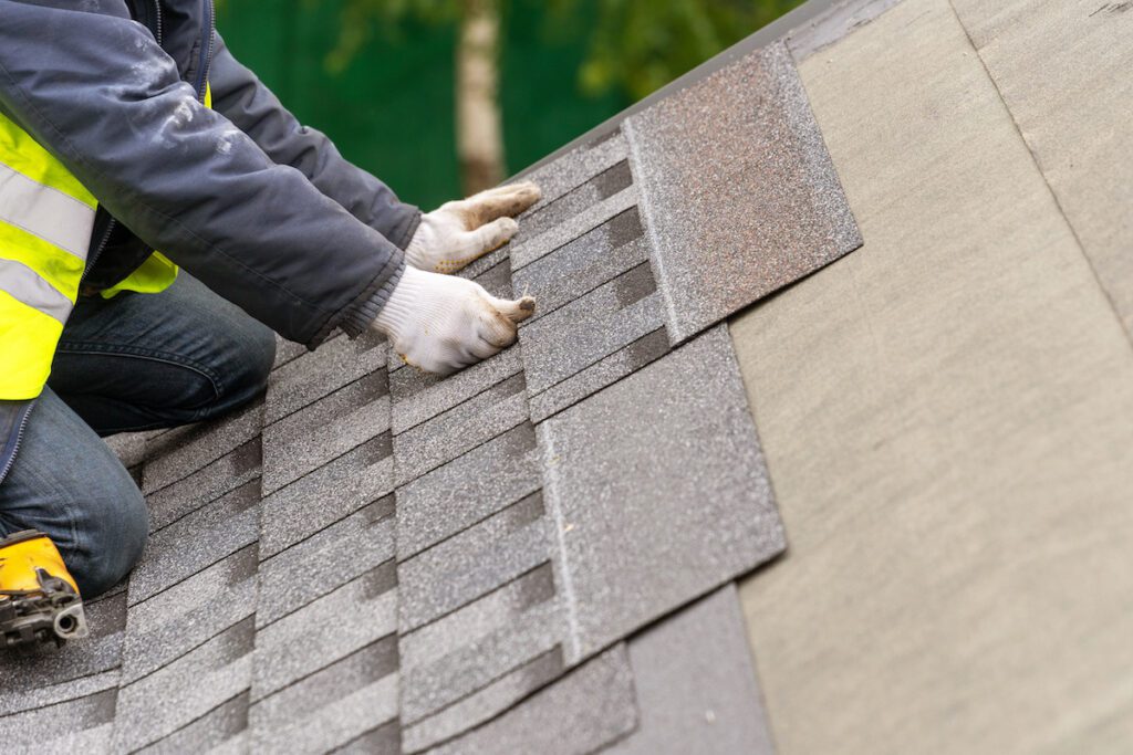 Close up and real photo of professional workman in special protective work wear holding in hands asphalt or bitumen tile and installing it on top of the roof under construction house