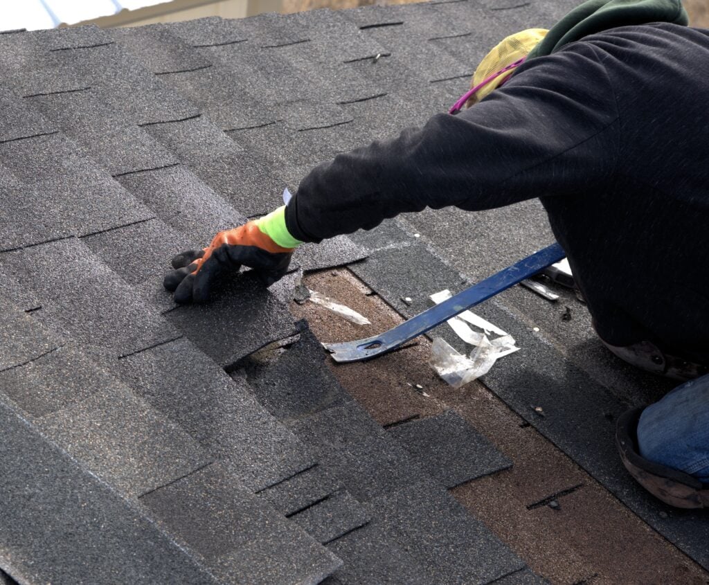 How To Find & Replace Damaged Roof Shingles - All Phase Roofing & Restoration Roofer uses a blue pry bar to remove damaged shinlges during the repair of a residential roof.