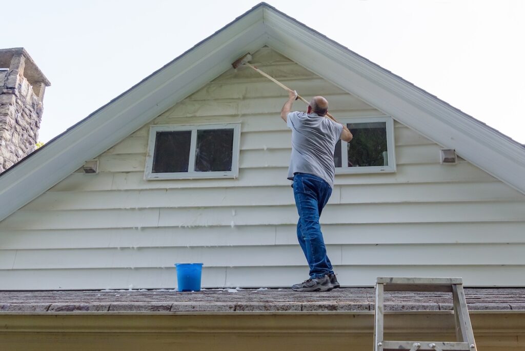 Soft Wash Roof Cleaning: Step-By-Step Guide - All Phase Roofing & Restoration Man stands on porch roof to wash house with brush on extension