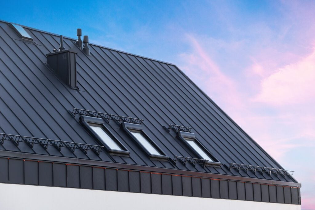 5 Signs You Need Metal Roof Replacement - All Phase Roofing & Restoration House with metal galvanized coating, chimney pipe, mansard windows and snow holding cornice on roof slope against sunset sky. Element of modern residential building with zinc rooftop sheets