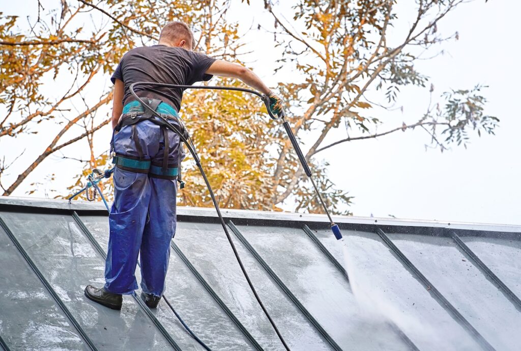 Commercial Roof Cleaning (7 Tips to Extend Lifespan) - All Phase Roofing & Restoration Man, cleaner with high pressure water sprayer wash steel dirty roof.Worker in waterproof uniform with pressure washer gun wash roof of private house. Roof cleaning with water pressure cleaner commercial roof cleaning
