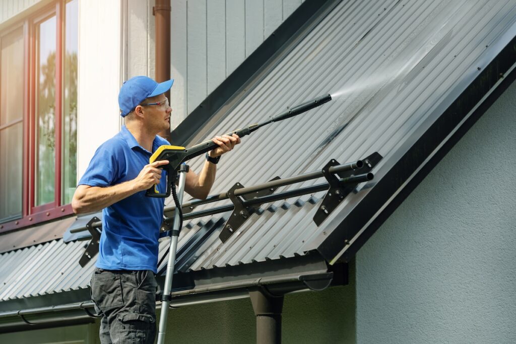 Commercial Roof Cleaning (7 Tips to Extend Lifespan) - All Phase Roofing & Restoration man standing on ladder and cleaning house metal roof with high pressure washer